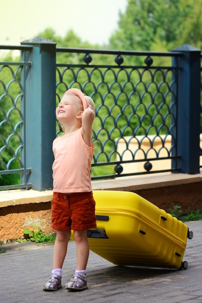 The little girl smiles and frowns in the sun. A child in a hat and red shorts is carrying a large yellow suitcase.