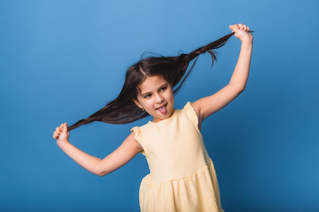 Little girl smiles and pulls her long braided hair  isolated on blue background