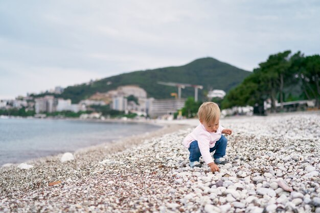 Little girl squatting on a pebble beach and holding a pebble in her hand