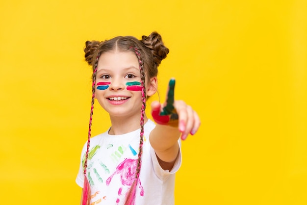 A little girl stained in multicolored paints for drawing stretches her index finger forward pointing at your recalama on a yellow isolated background The development of art and children's creativity