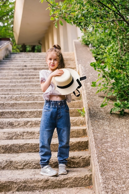 A little girl stands in the park and holds a hat in her hands. The child is happy and laughing.