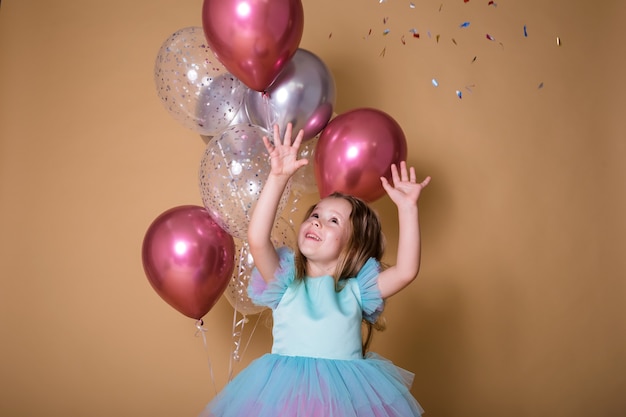 Little girl stands with a bunch of inflatable balloons catching confetti on a beige background with a place for text