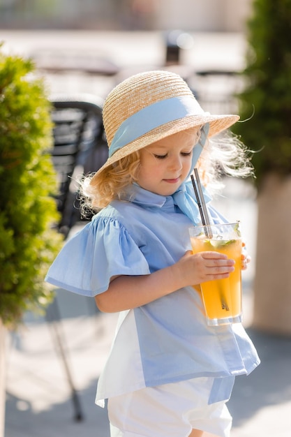 little girl in a straw hat and a blue dress drinks juice from a glass outdoors