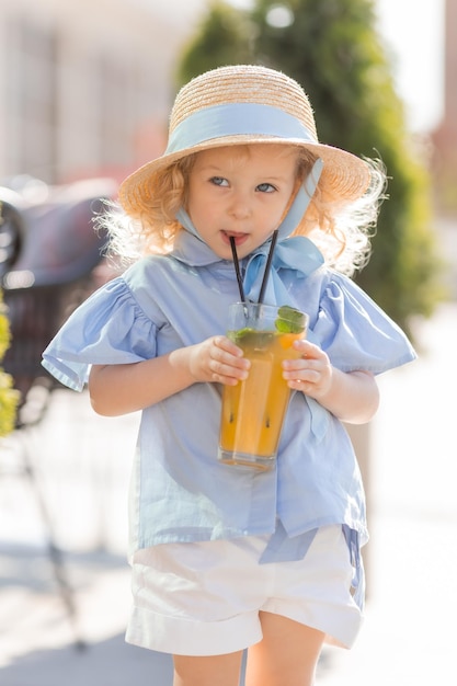 little girl in a straw hat and a blue dress drinks juice from a glass outdoors