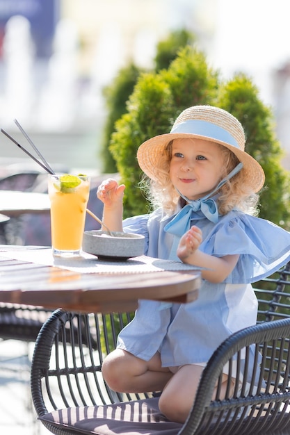 little girl in a straw hat and a blue dress is having breakfast outdoors