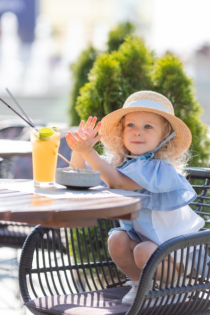 little girl in a straw hat and a blue dress is having breakfast outdoors