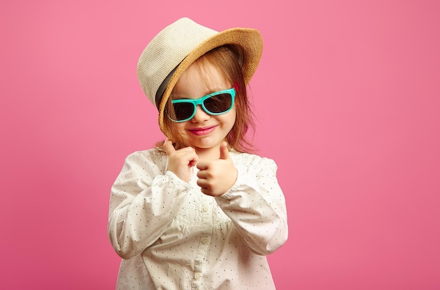 Little girl in straw hat and sunglasses shows a thumb up glances at you stands on pink isolated