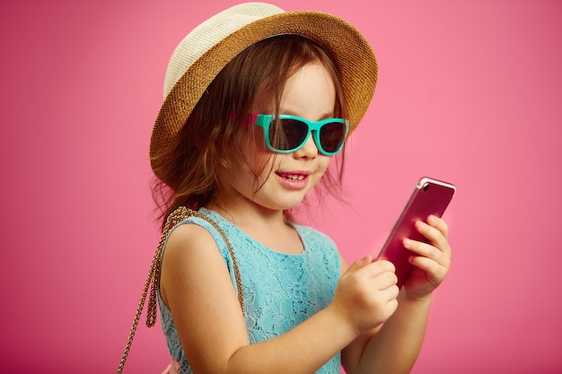 Little girl in summer clothes wearing a beach hat sunglasses looking at the phone stands on pink