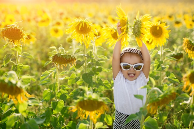 A little girl in sunglasses laughs while holding two flowers in a huge field of sunflowers at sunset