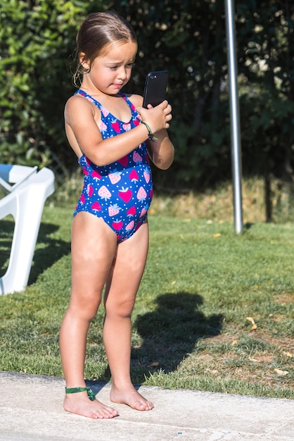 Little girl in a swimsuit standing on the edge of the pool looking at the mobile phone Childhood children smartphone technology vacation internet and fun concept