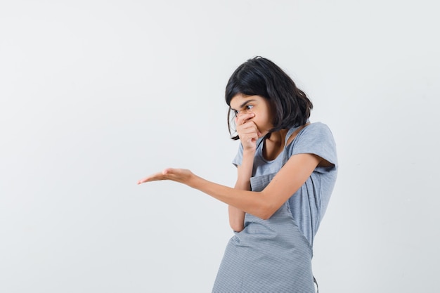 Little girl in t-shirt, apron stretching hand in questioning manner and looking surprised , front view.