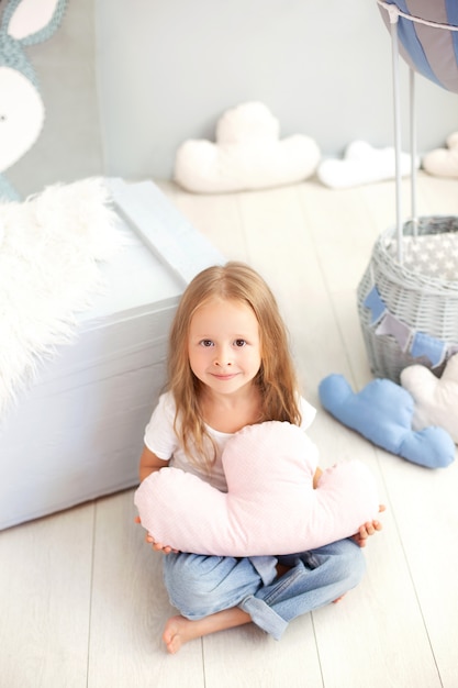 A little girl in a T-shirt and jeans holds a cloud pillow against the wall of a decorative balloon. Toddler is playing in the children's room. The concept of childhood, travel. Interior room