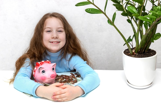 Little girl at the table happily hugs a piggy bank and money savings financial literacy