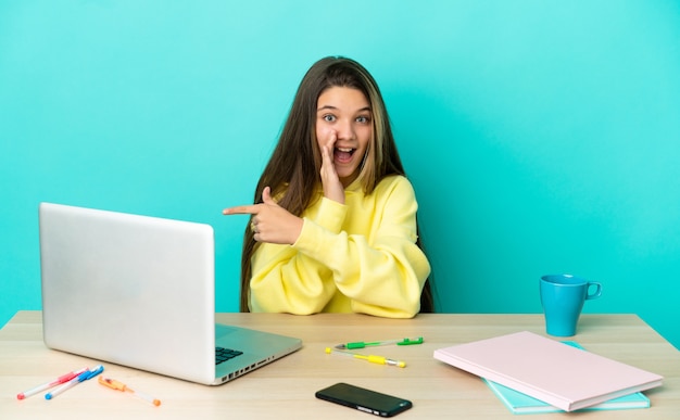 Little girl in a table with a laptop over isolated blue background pointing to the side to present a product and whispering something