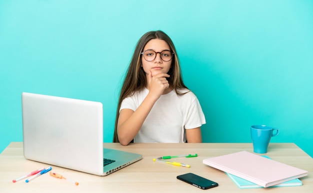 Little girl in a table with a laptop over isolated blue background thinking