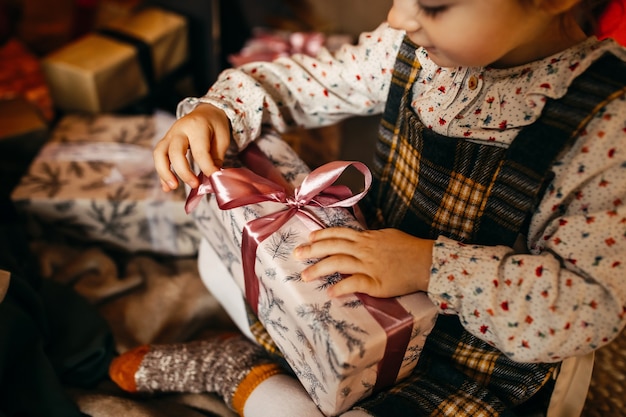 Little girl unwrapping gift, sitting next to a Christmas tree