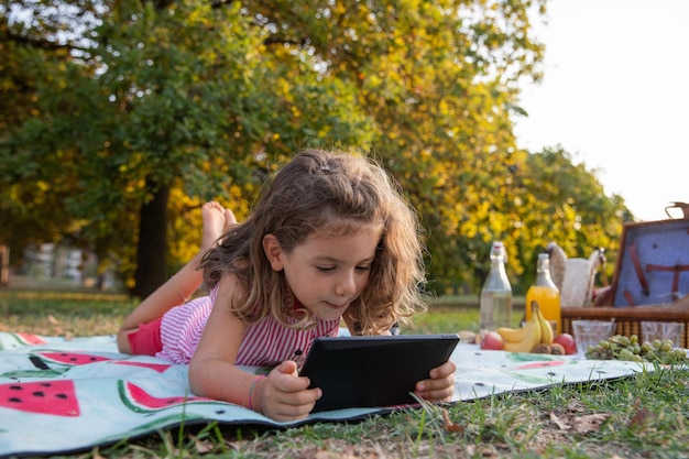 A little girl uses a tablet while on a picnic in a park children and education with technology