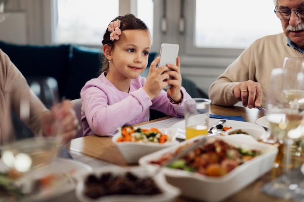 Little girl using cell phone while having lunch with her grandparents at dining table