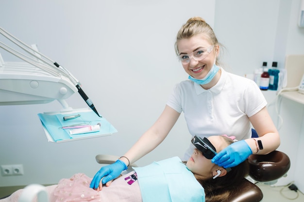 Little girl in VR glasses and wireless headphones sitting in dental chair Dentist with assistant