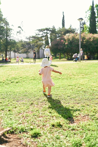 Little girl walks along a green lawn in the park to people walking in the distance