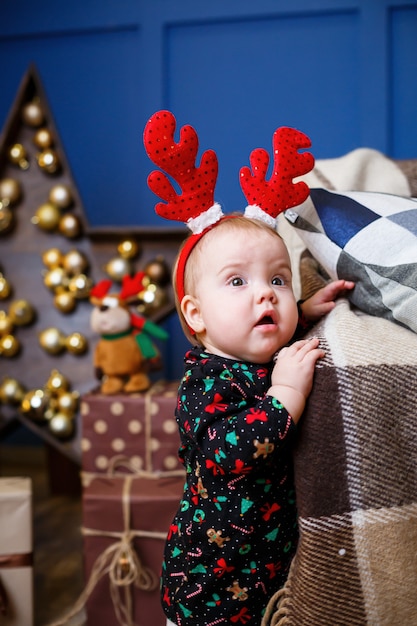 A little girl in warm sweater sits under a Christmas tree with toys and gifts with horns on her head. Happy childhood. New Year holiday atmosphere