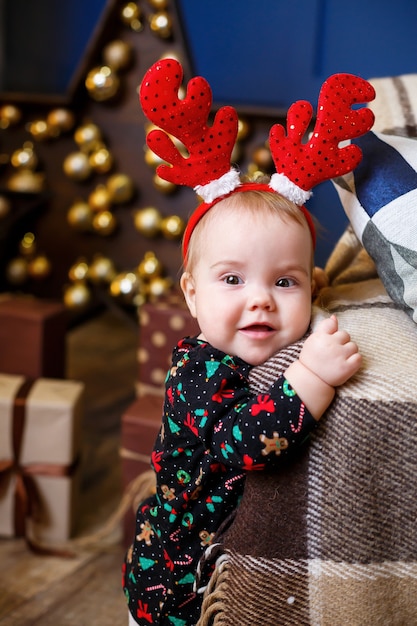 A little girl in warm sweater sits under a Christmas tree with toys and gifts with horns on her head. Happy childhood. New Year holiday atmosphere