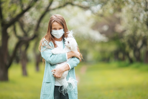 Little girl wearing a protective mask is walking alone with a dog outdoors because of the coronavirus pandemic