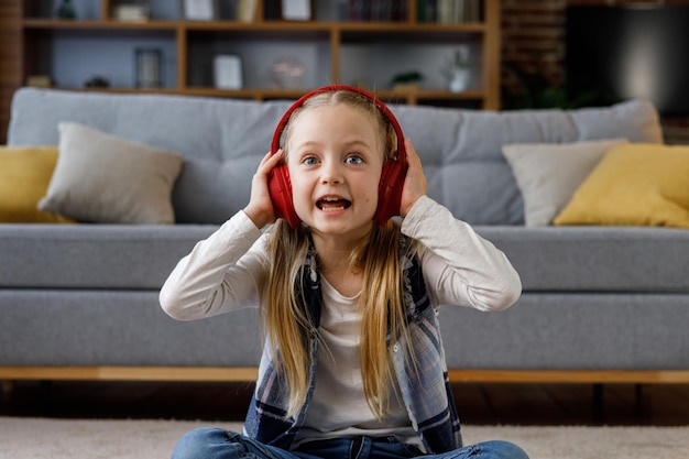 Little girl wearing red earphones looking at camera Portrait of cute smiling child resting indoors at home interior Concept of a happy childhood