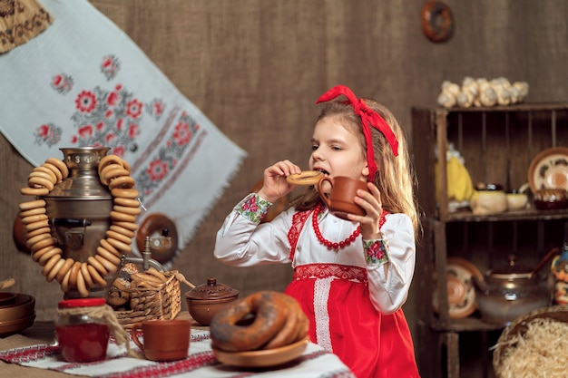 Little girl wearing red headband and ornamental shirt drinking tea from samovar celebrating Maslenitsa