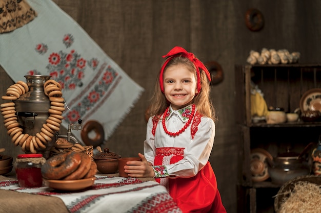 Little girl wearing red headband and ornamental shirt holding cup of tea from samovar celebrating Maslenitsa