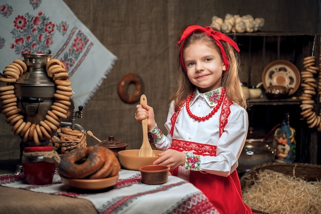 Little girl wearing red headband and ornamental shirt sitting at table full of food and big samovar celebrating Maslenitsa