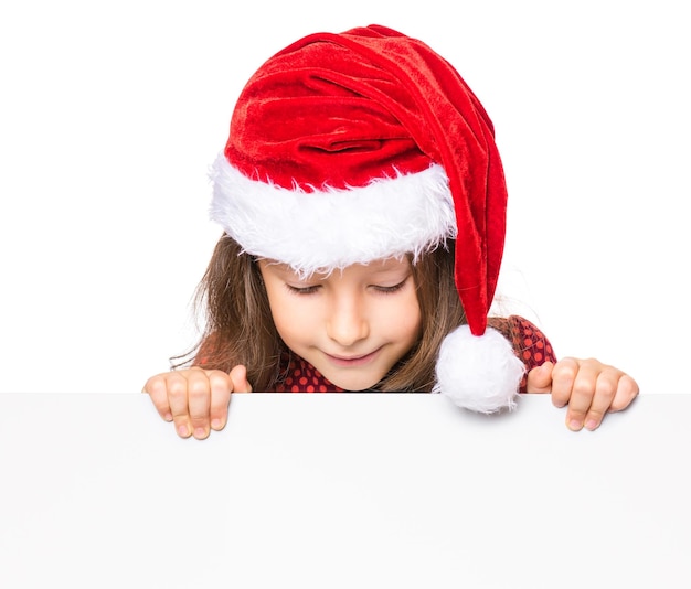 Little girl wearing Santa Claus hat posing behind white panel isolated on white background Kid holding empty Christmas billboard Child peeping behind blank holidays board