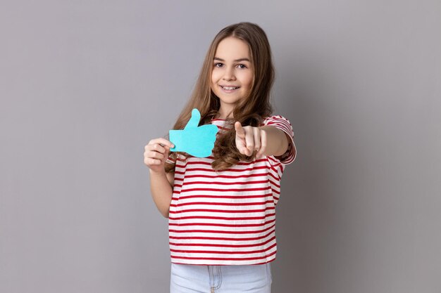 Little girl wearing Tshirt holding blue thumbs up sign pointing finger at you asking to like posts