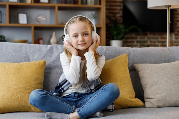 Little girl wearing white earphones looking at camera Portrait of cute smiling child resting indoors at home interior Concept of a happy childhood