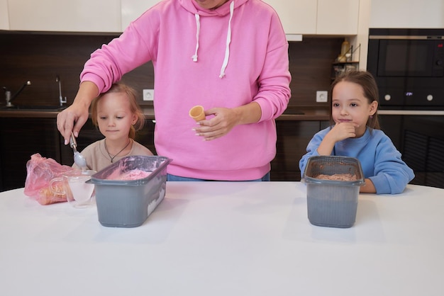 Little girl in a white blouse happily eats ice cream from a snail at a table in the kitchen