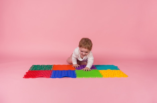 A little girl in a white bodysuit crawls on all fours on an orthopedic Mat