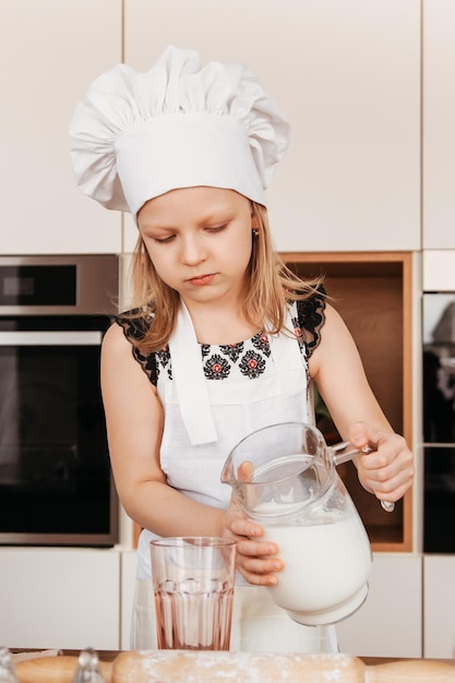 A little girl in a white chef's hat pours milk from a jug into a glass in the kitchen