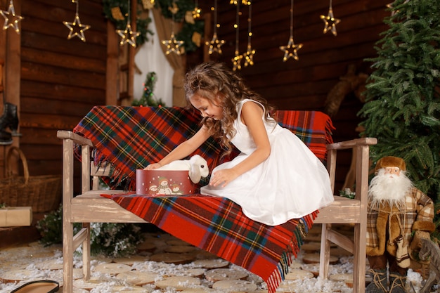  A little girl in white dress sitting on the bench and open box with white rabbit. Magic Light in Night Xmas Tree Interior