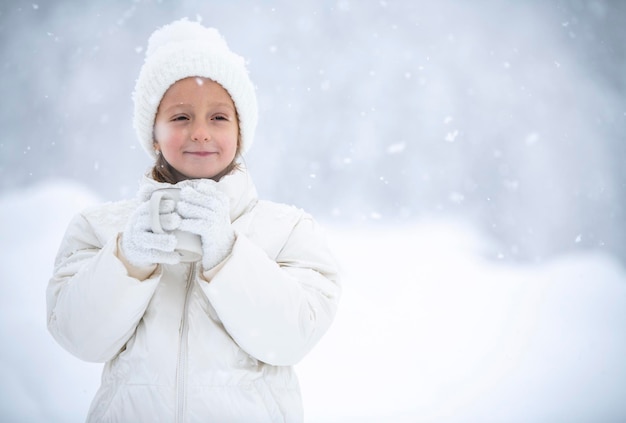 A little girl in a white jacket and a white hat is holding a cup of tea in her hands during a snowfall against the backdrop of beautiful snowdrifts and forests