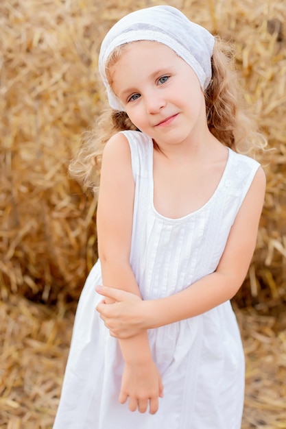 a little girl in a white scarf and sundress stands against the background of a haystack summer