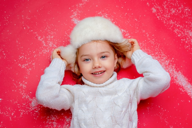 a little girl in a winter hat is lying on the floor in the snow on a red background