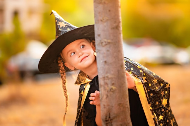 little girl in a witch costume celebrating Halloween in the park peeking out from behind a tree