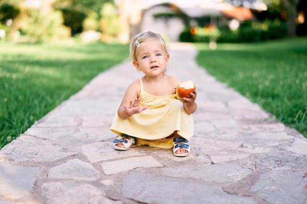 Little girl with apple in hand sits on a cobbled path in the park