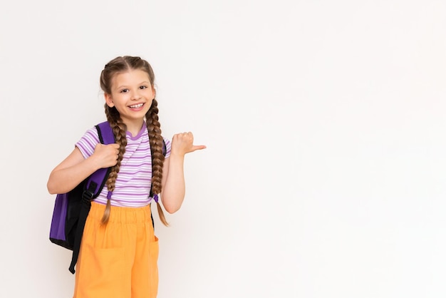 A little girl with a backpack on her back points sideways with her thumb at your advertisement on a white isolated background Courses for schoolchildren summer education for children