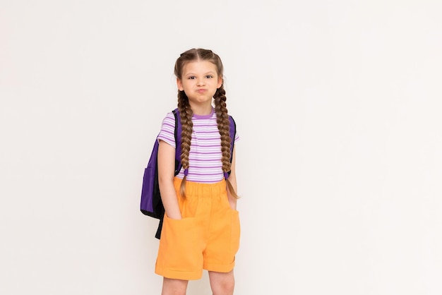 A little girl with a backpack on her back puffed out her cheeks and put her hands in the pockets of her shorts on a white isolated background