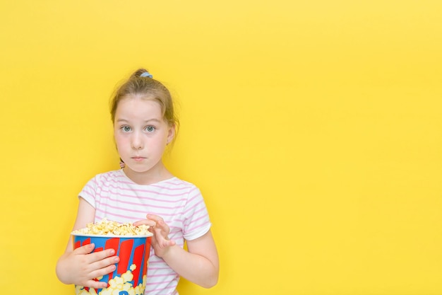 Little girl with big eyes in surprise with a glass of popcorn on a yellow background with copy space