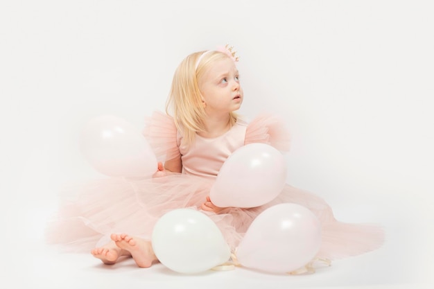 Little girl with blonde hair in lush dress with balloons in hands sits on floor isolated on white background