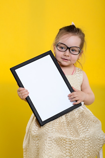 Little girl with board in beautiful dress and glasses on yellow background