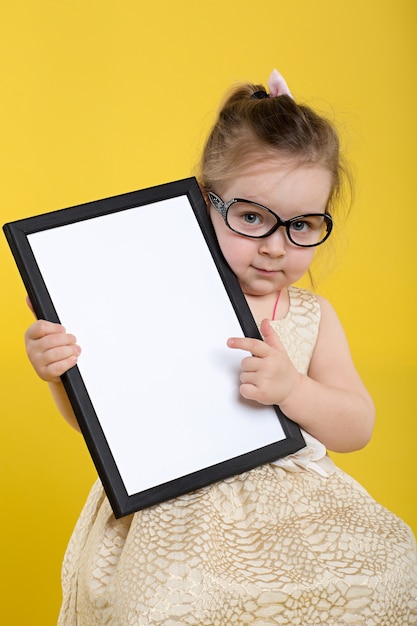 Little girl with board in beautiful dress and glasses on yellow background