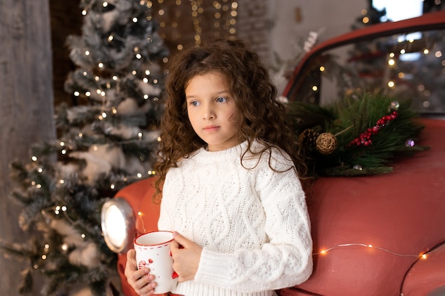 Little girl with Christmas cup of tee near red car and Christmas trees with lights. Merry Christmas and Happy Holidays